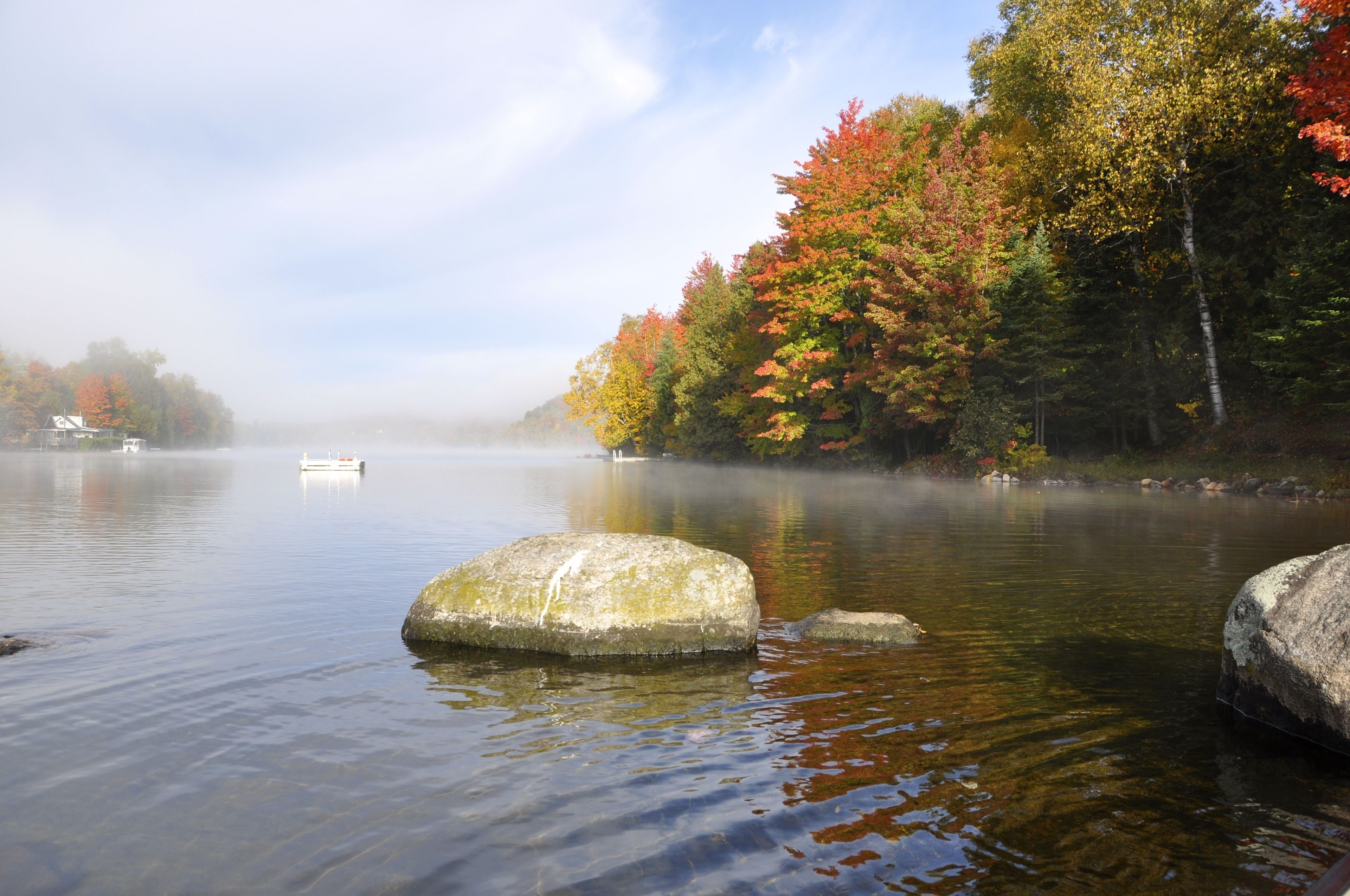 Un lac paisible, avec de gros rochers au premier plan, entouré d’arbres aux feuilles d’automne.