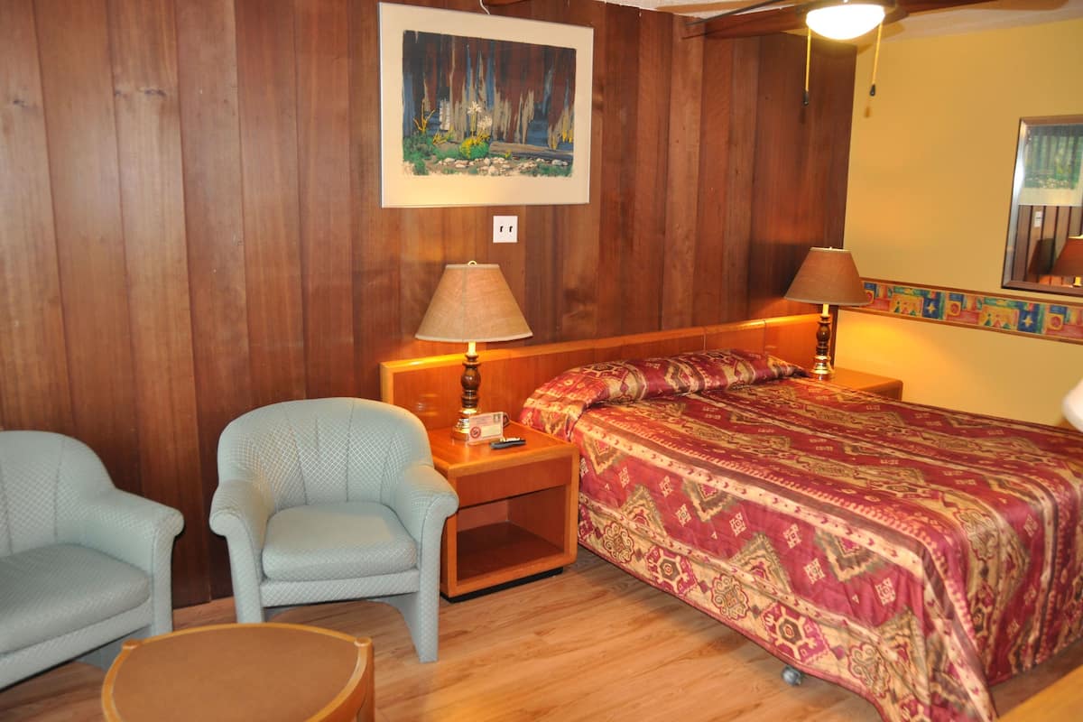Una habitación con pared de paneles de madera, una cama con colcha estampada, dos sillas, una mesita y un cuadro sobre la cama.