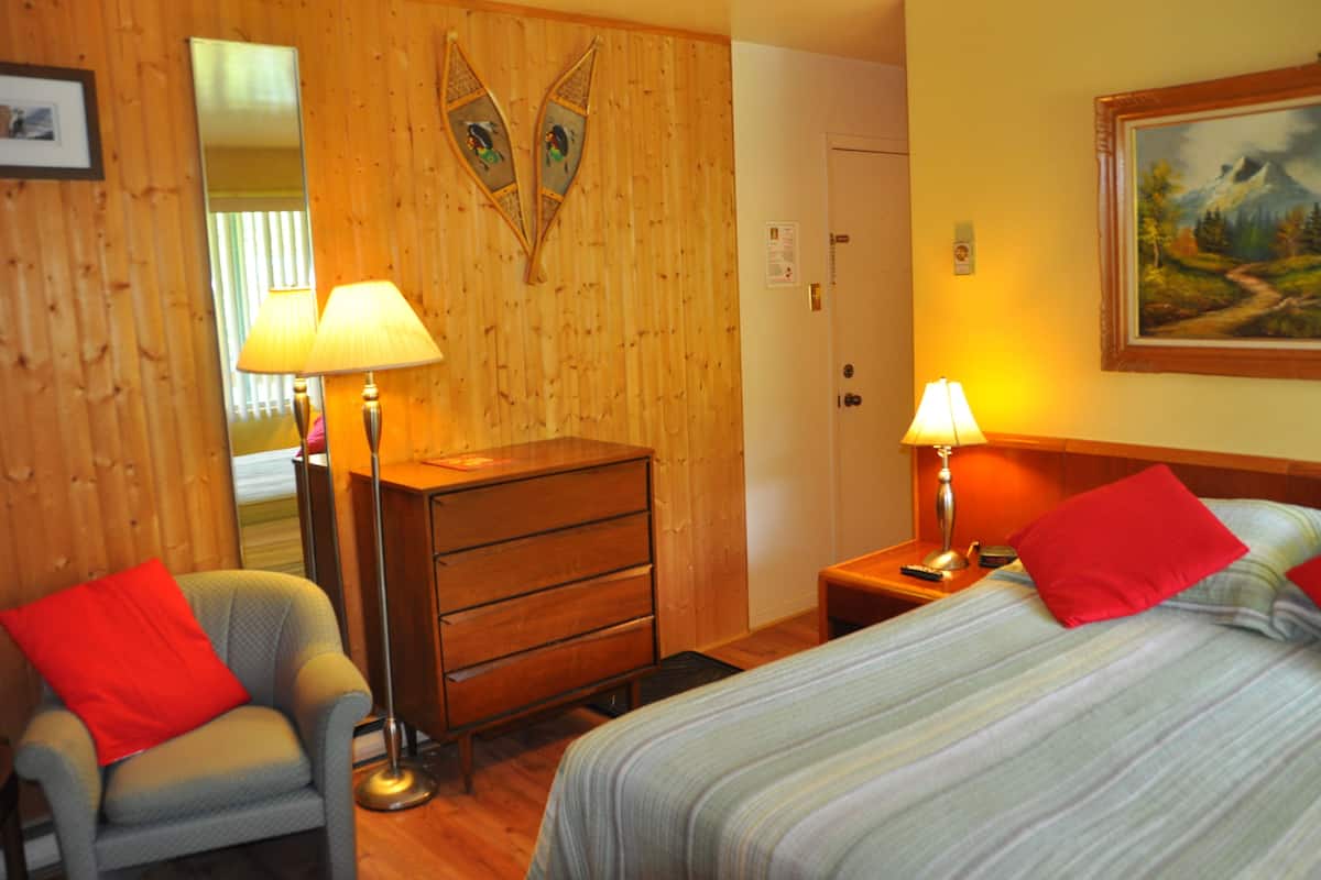 Habitación de hotel con paredes de paneles de madera, una cama con ropa de cama a rayas, una mesita de noche con una lámpara, un aparador y una silla con un cojín rojo.