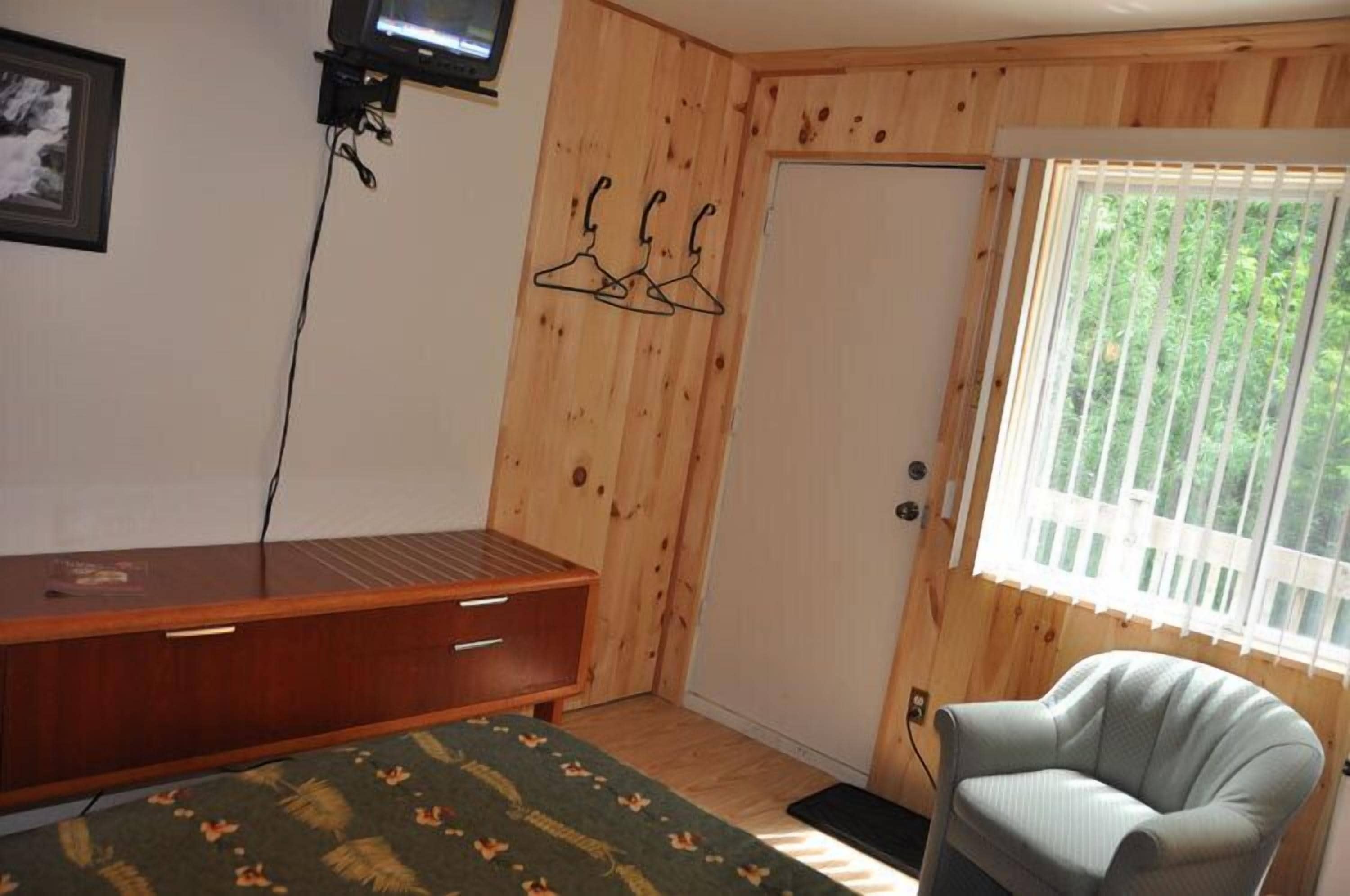 A room with wooden walls, a bed with a green patterned bedspread, a wooden desk with drawers, a chair, and a window with blinds.