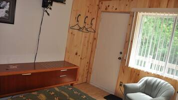 A room with wooden walls, a bed with a green patterned bedspread, a wooden desk with drawers, a chair, and a window with blinds.