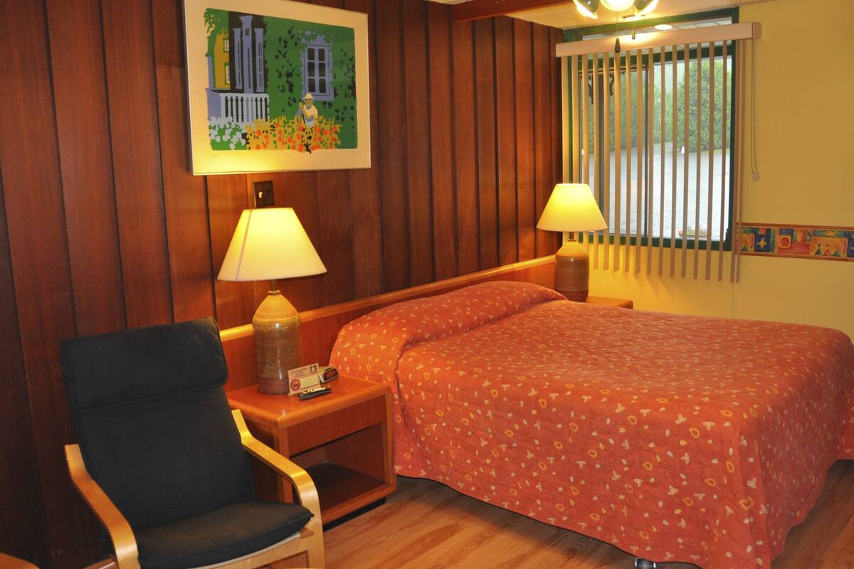 Una habitación con cama, una silla, una mesita de noche, una lámpara y un cuadro en la pared.