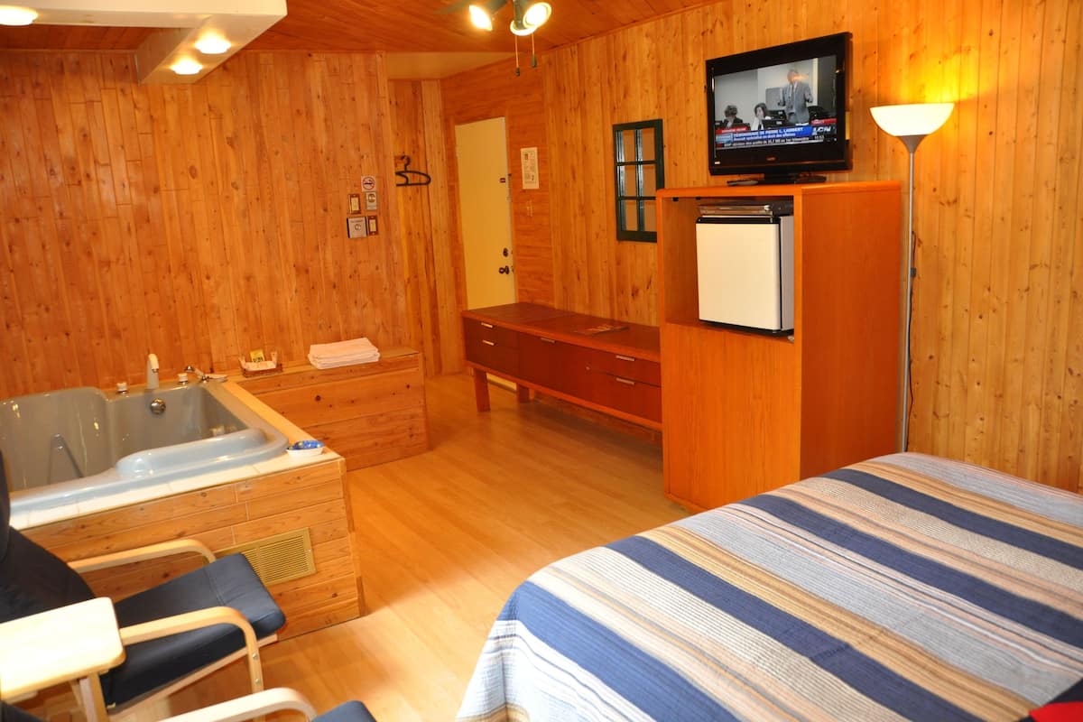 Una habitación de cabaña de madera con una cama, un escritorio pequeño, una silla, un televisor y un baño con bañera y lavabo.
