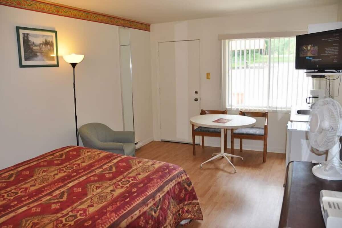 Habitación de hotel con cama, escritorio, silla, mesa, televisor y ventana con persianas.