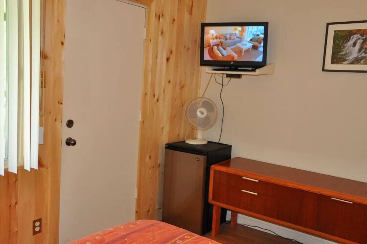 Una habitación con puerta de madera, televisor montado en la pared, un pequeño mueble de madera y un banco de madera.