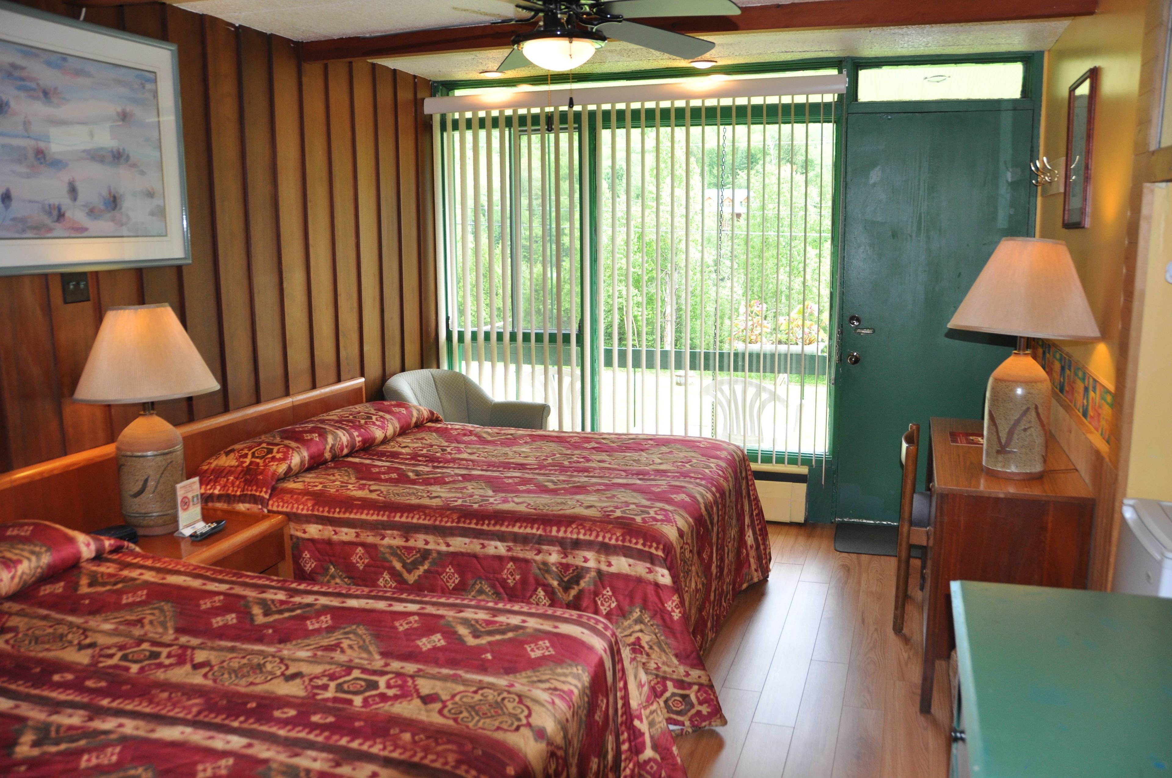 A room with two beds, a ceiling fan, a door, and a window with blinds.