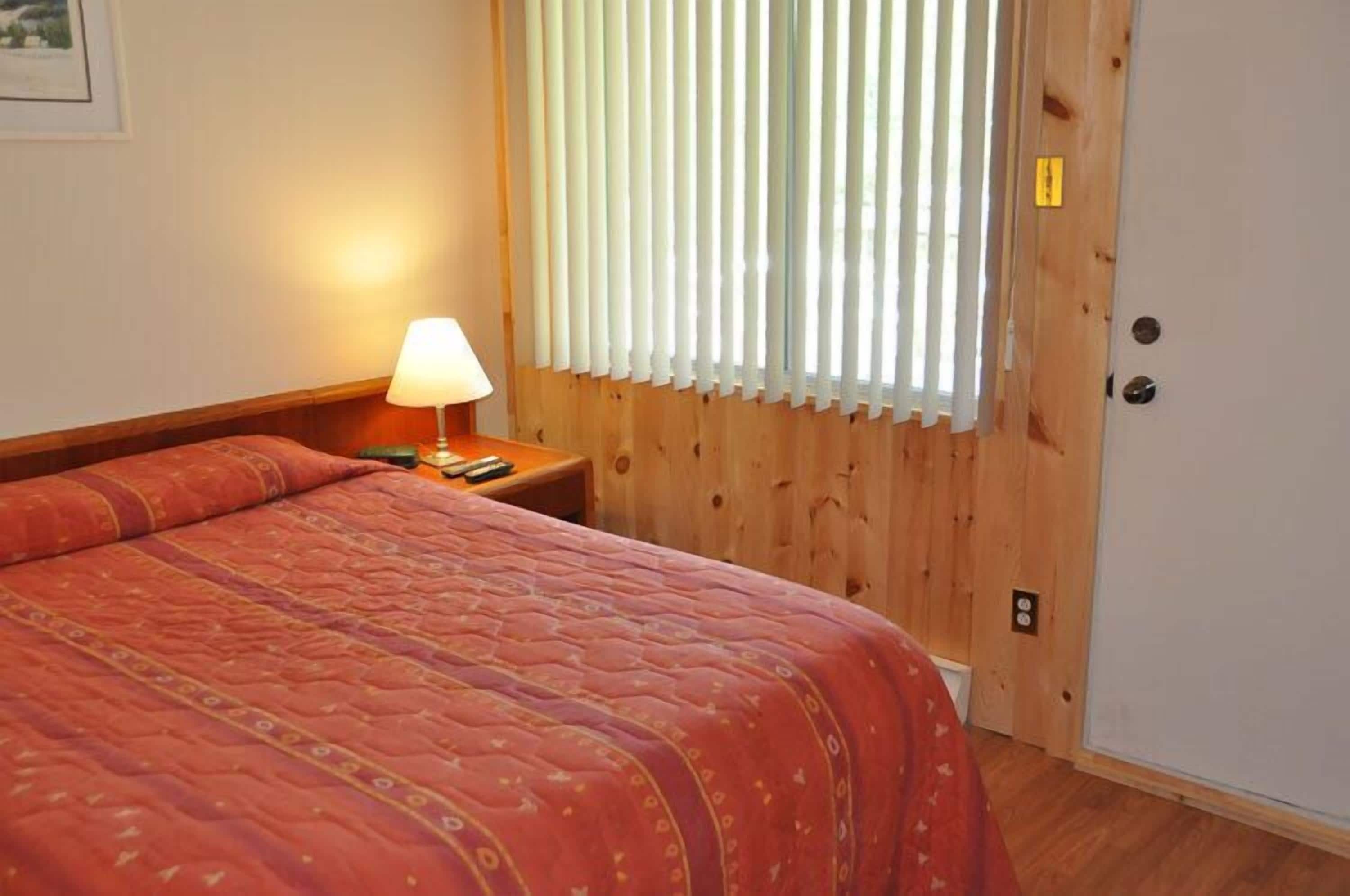 Una habitación con una cama, una mesita de noche, una lámpara y una ventana con persianas.