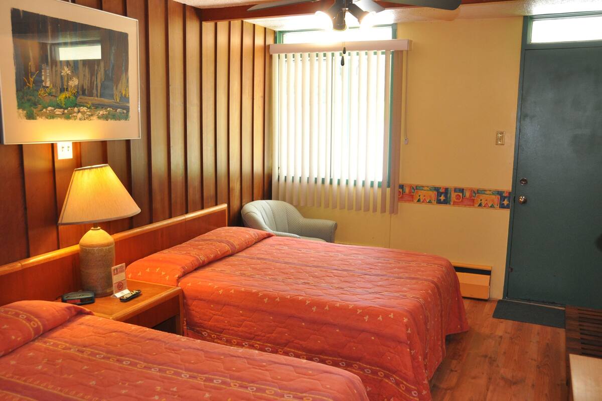 Habitación de hotel con dos camas, una mesita de noche con lámpara, una silla, una ventana con persianas y una puerta.