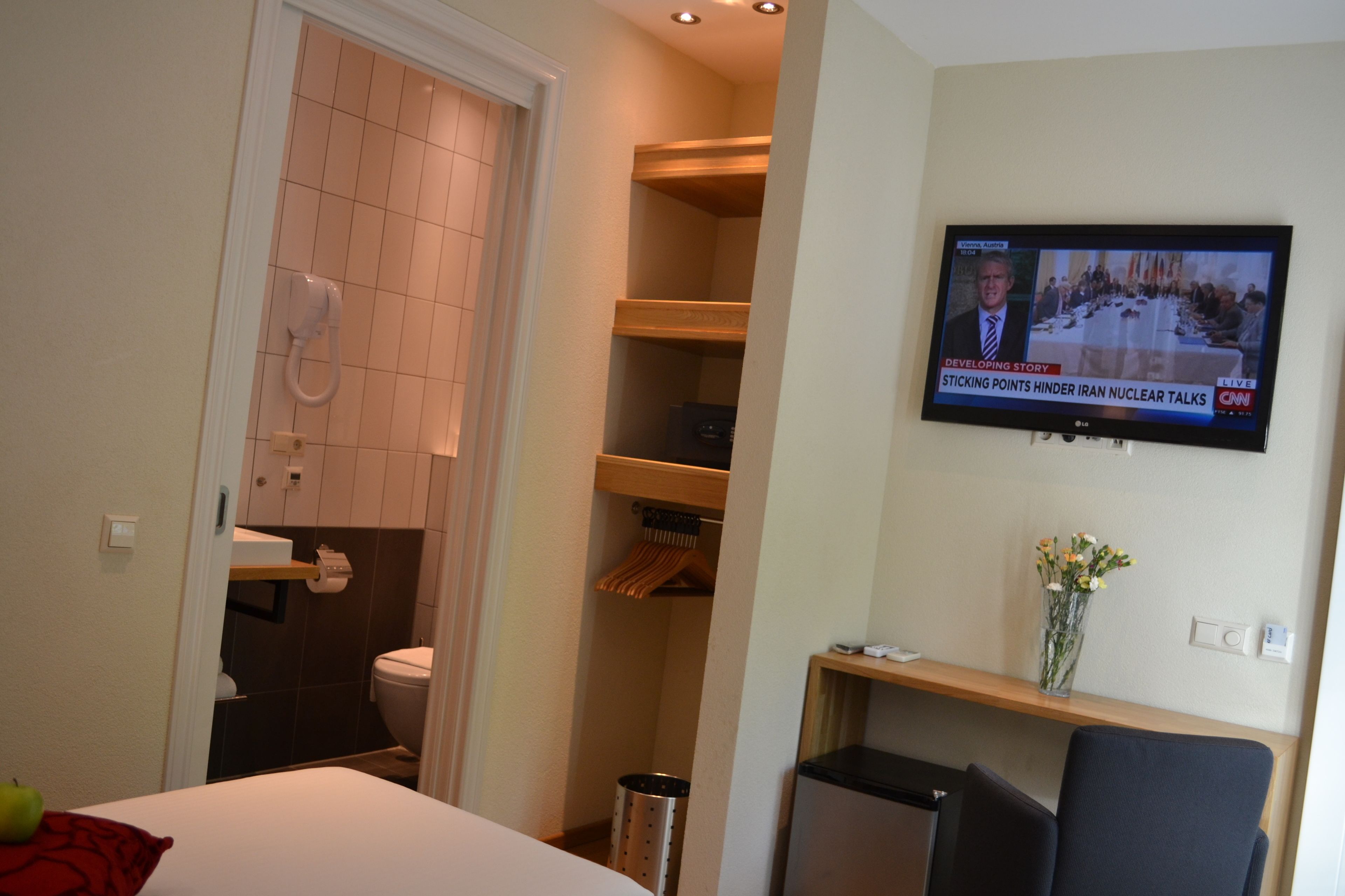 A hotel room with a television displaying a news program, a bed, a small table with a vase, and a closet with shelves.