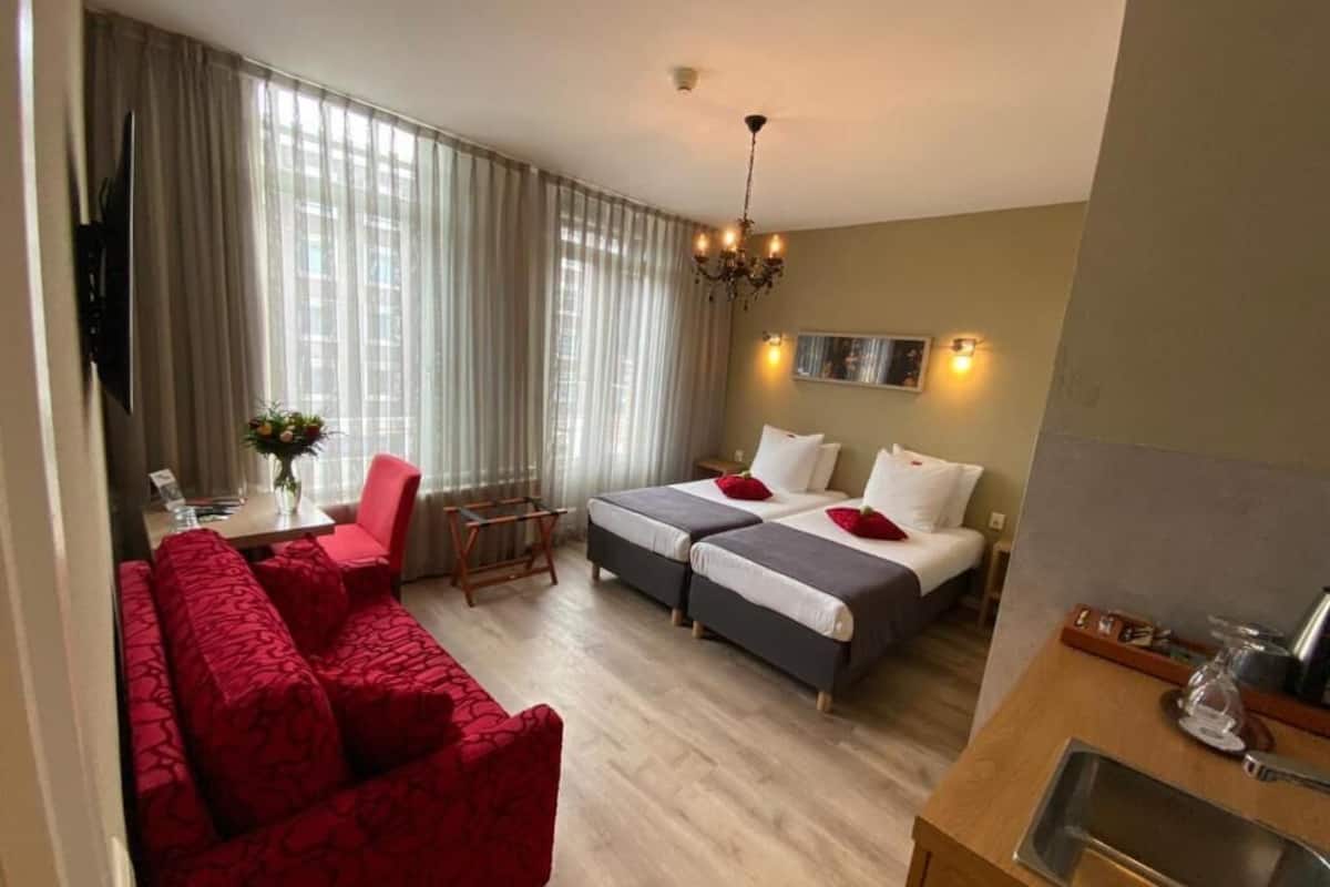 A hotel room with a red sofa, two single beds, a dining area with a table and chairs, a chandelier, and a large window with curtains.