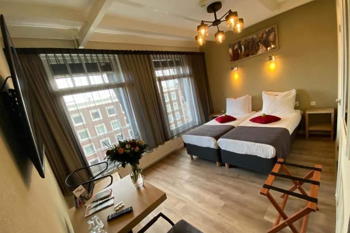 A hotel room with a desk, two beds, a TV, and a large window with curtains.