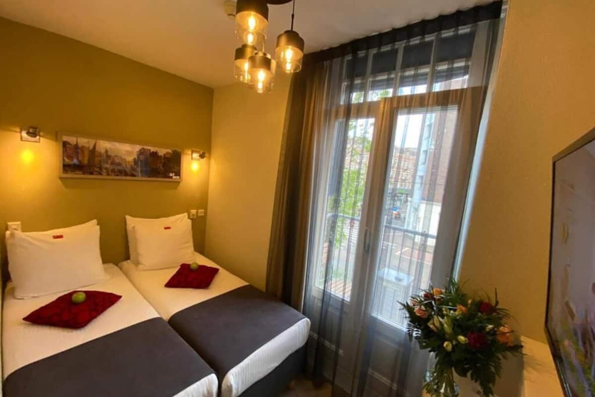 A hotel room with two beds, a window with sheer curtains, a hanging light fixture, and a painting on the wall.
