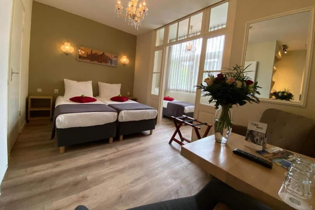 A hotel room with two beds, a wooden floor, a chandelier, a desk with a vase of flowers, and a large mirror.