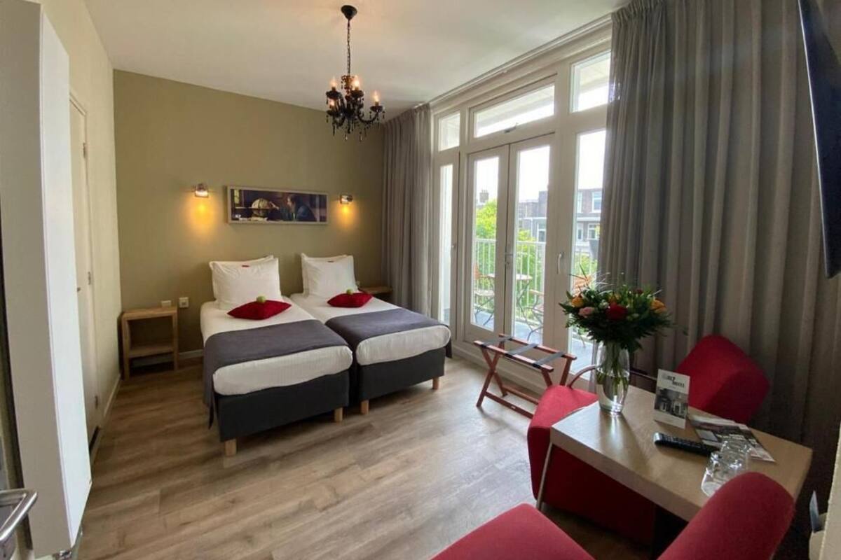 A hotel room with two beds, a desk, a chair, a chandelier, and large windows with curtains.