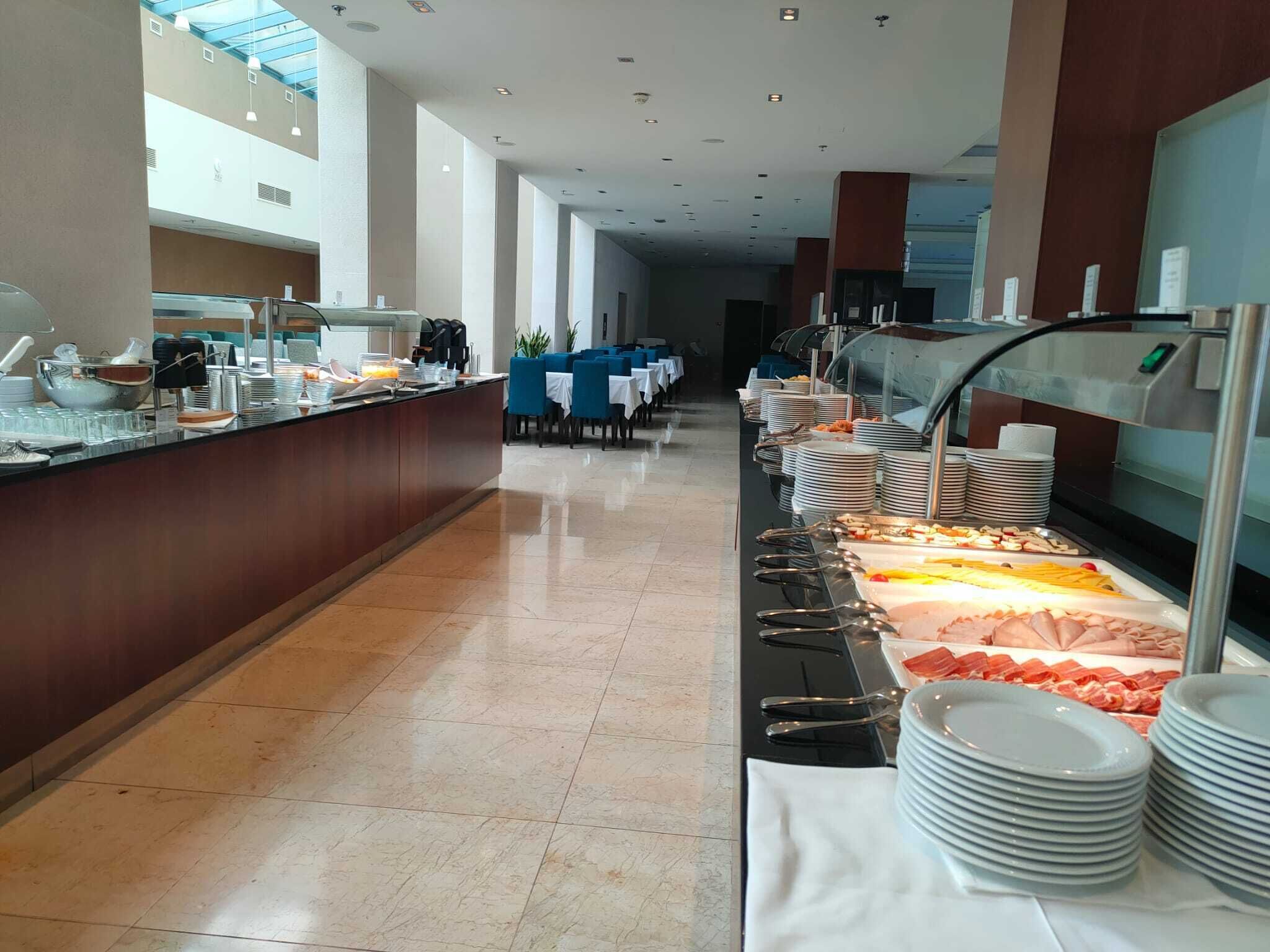 Montaje tipo bufé con diversos alimentos y utensilios, una larga barra con asientos y vista del comedor al fondo.