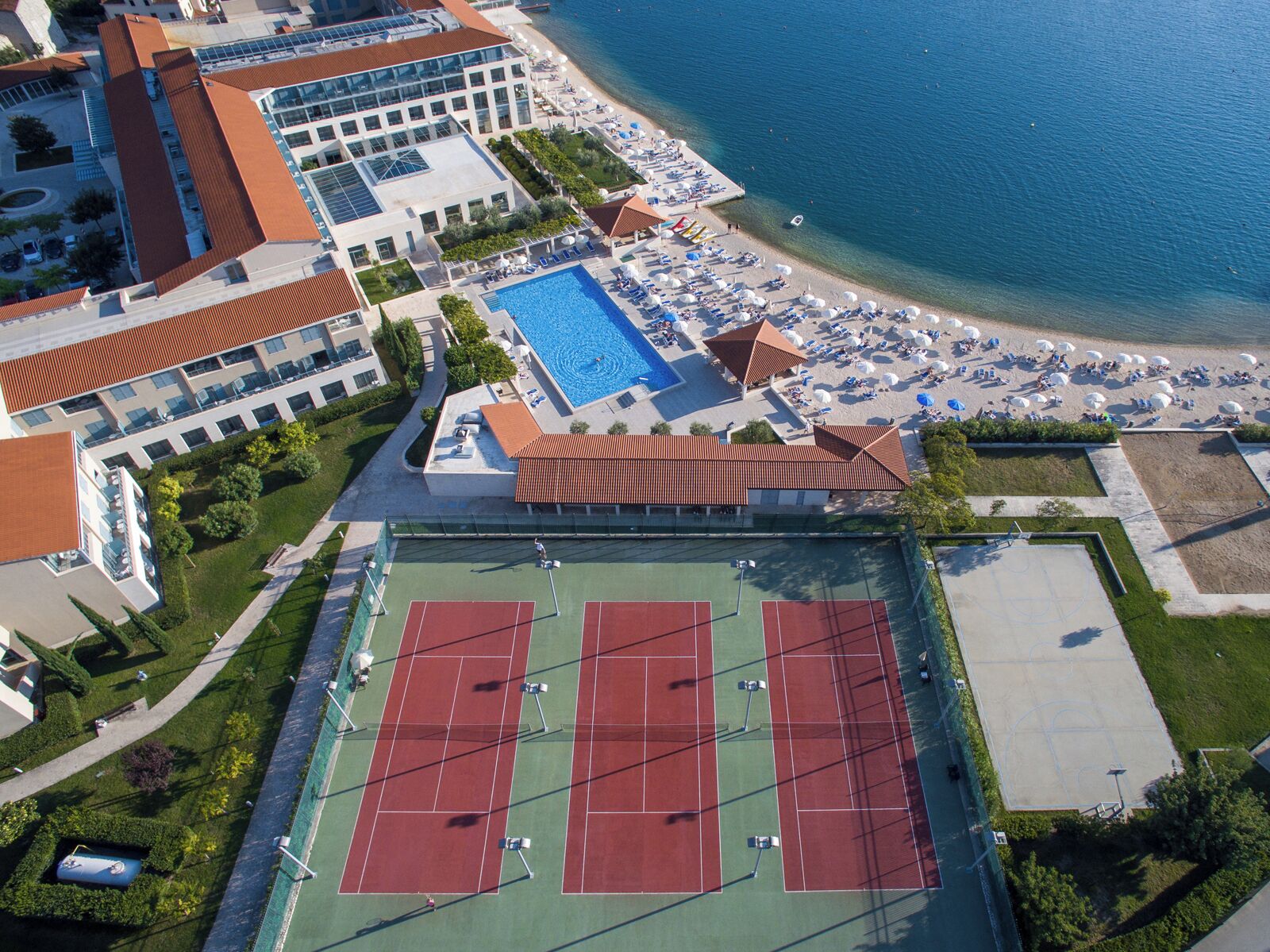 Vista aèria d'un hotel a la costa, amb piscina, platja, pistes de tennis i zona de pàrquing.