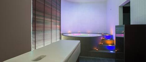 A modern spa room with a massage table, a hot tub, and a small seating area.