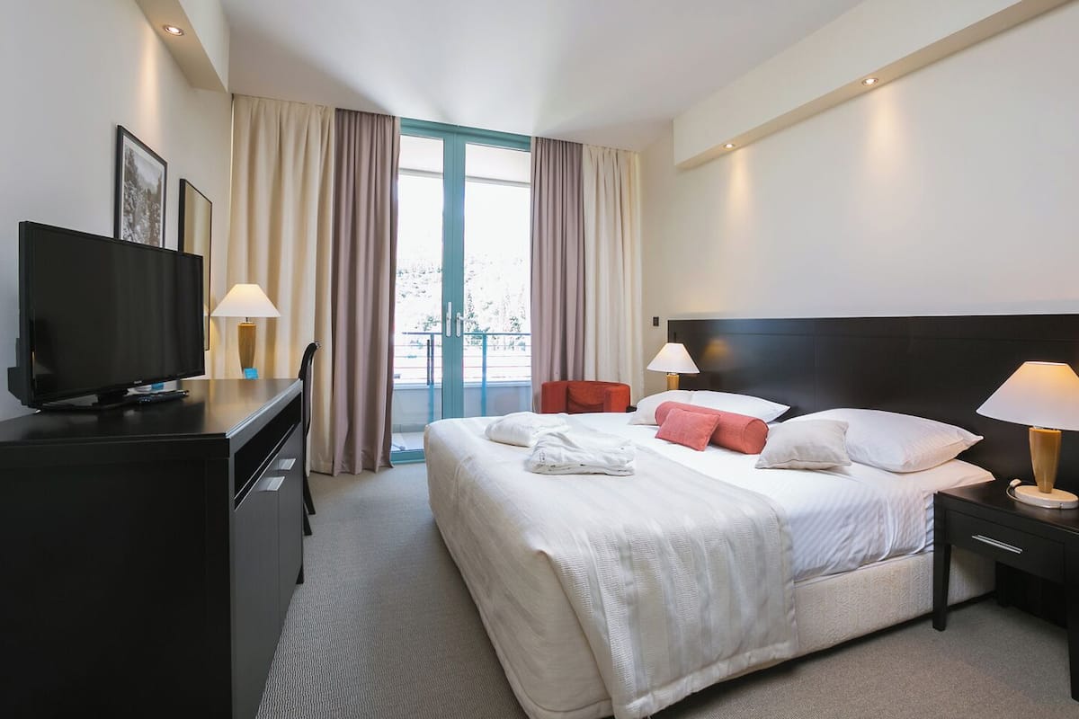 Une chambre d’hôtel avec un grand lit, deux tables de chevet avec des lampes, un téléviseur posé sur une commode et une vue sur l’extérieur grâce à une grande fenêtre.