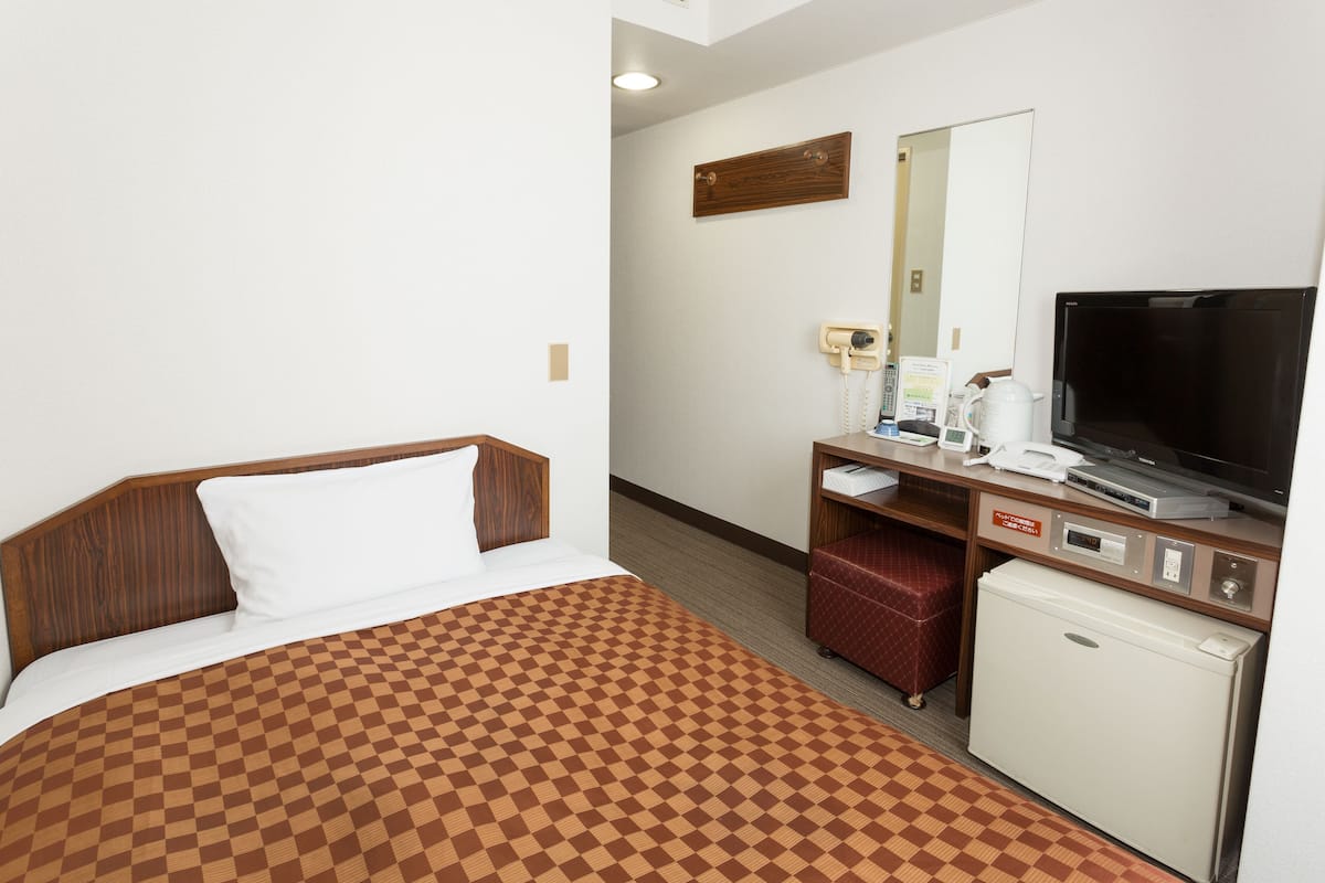 Habitación de hotel con colcha a cuadros, televisor, refrigerador pequeño, escritorio con espejo y banca tapizada de rojo.