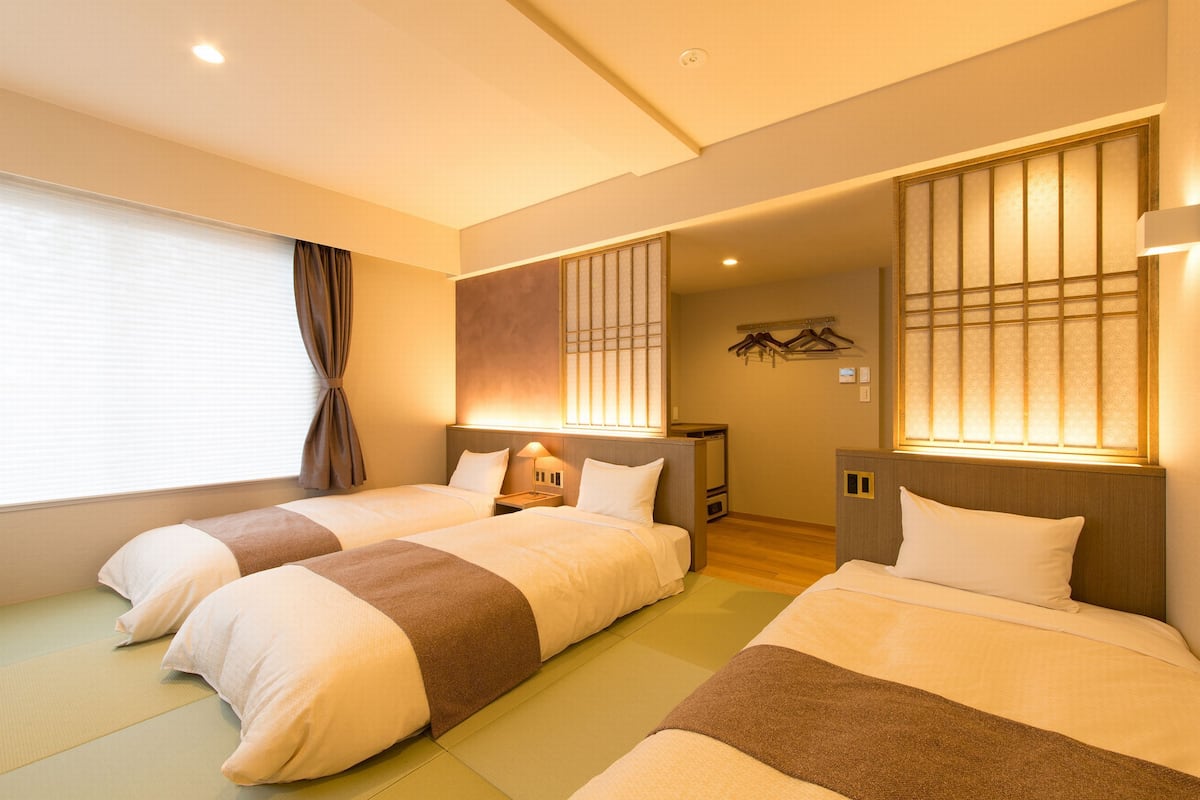 Una habitación de hotel con dos camas, un ventanal con cortinas, un televisor montado en la pared y una mesita de noche con lámpara.