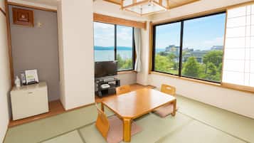 A room with a wooden table, chairs, a television, a small refrigerator, and large windows offering a view of greenery and buildings.