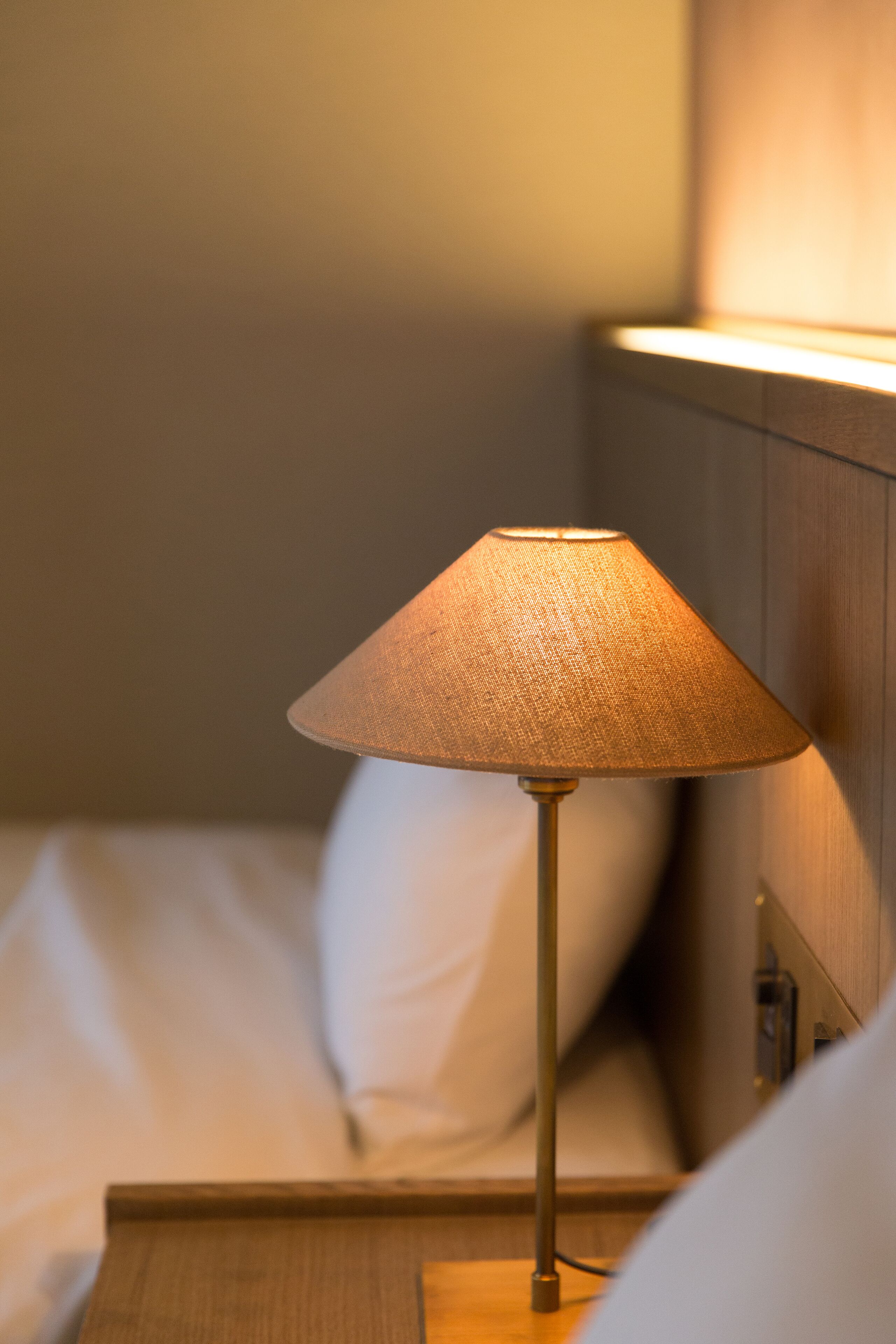 A lamp with a conical shade on a bedside table next to a bed.