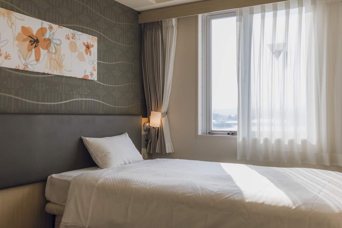 Habitación de hotel con una cama grande, cabecero con diseño floral, ventana con cortinas traslúcidas y lámpara de pared.