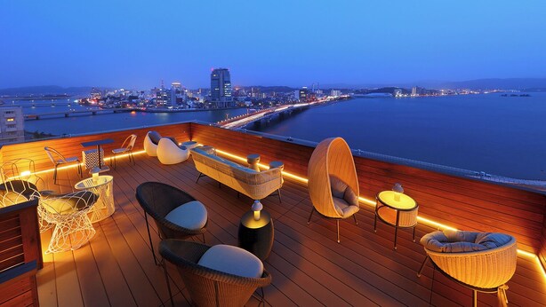 Una terraza en la azotea con mobiliario de mimbre, vistas a un horizonte urbano y al frente marítimo.