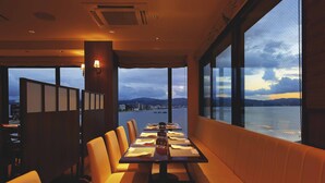 Un restaurante con una larga mesa dispuesta para comer, ventanas amplias que ofrecen vista a un cuerpo de agua y montañas distantes, y un separador con diseño tradicional.