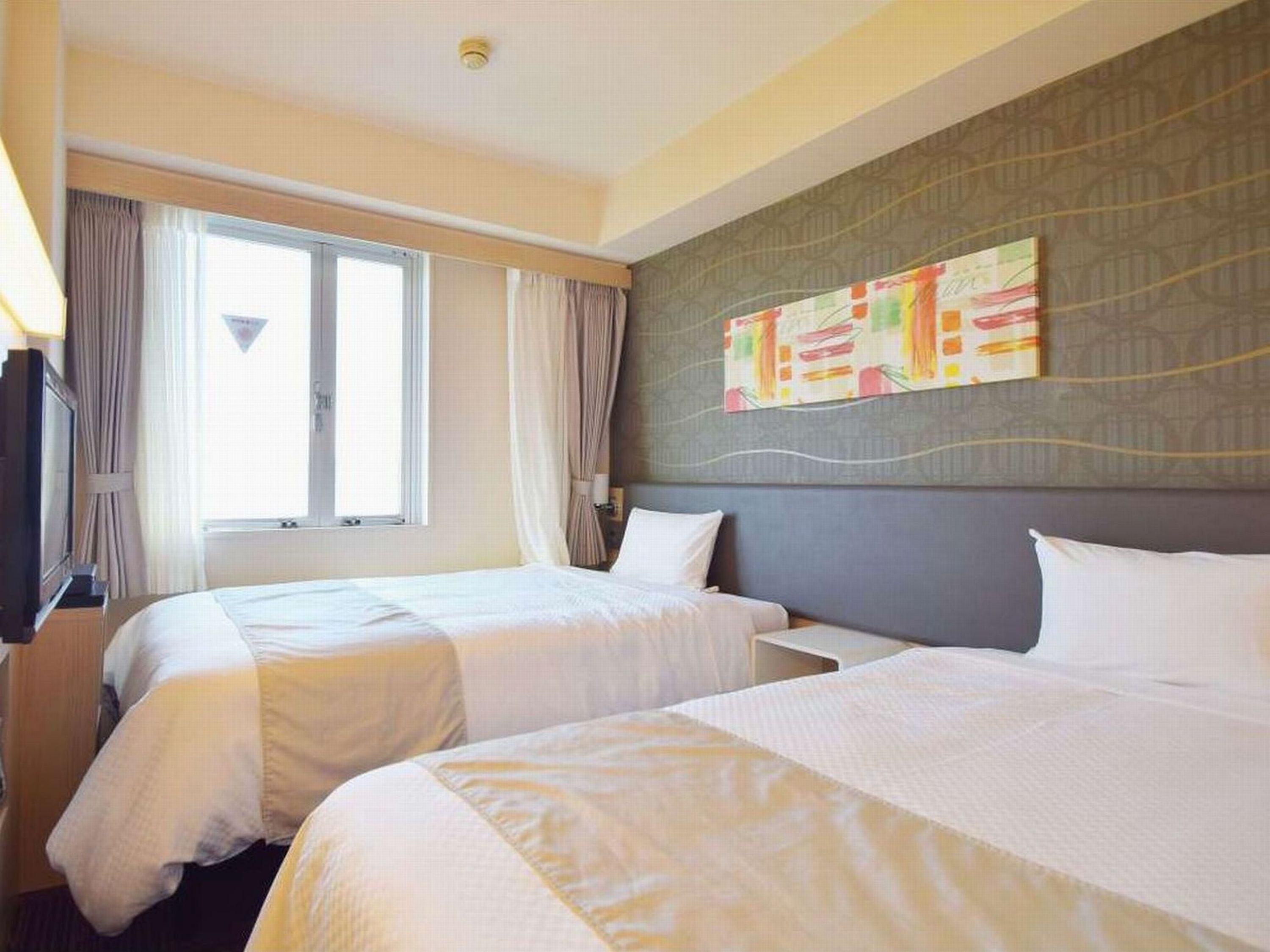 A hotel room with two beds, a large window with curtains, a flat-screen TV, and a wall with a colorful abstract painting.
