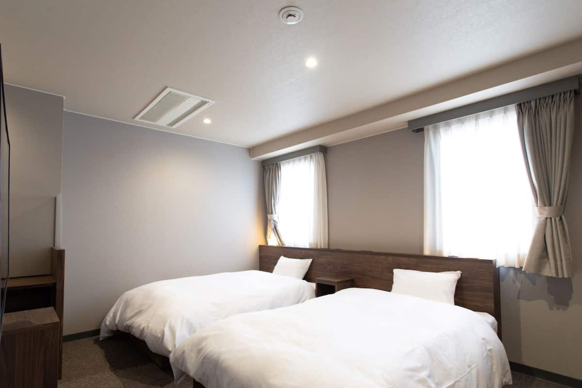 Habitación de hotel con dos camas individuales, cabecera de madera, mesita de noche pequeña, televisor integrado y ventanas grandes con cortinas.