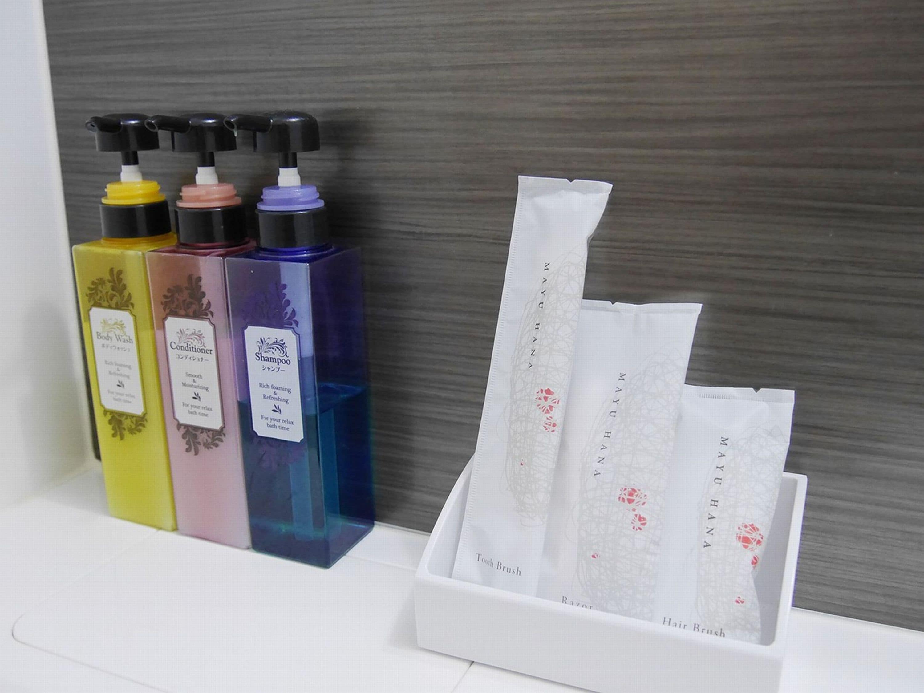 Un baño con un lavamanos blanco, tres botellas de champú, acondicionador y jabón de cuerpo, y un juego de maquinillas de afeitar y cepillos de pelo.