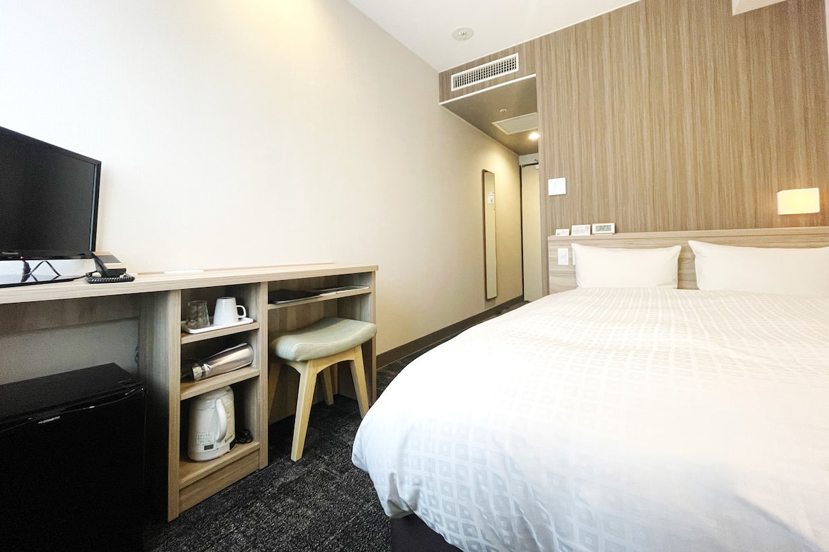 Una habitación de hotel con una cama, un escritorio con televisor, una silla y un estante con hervidor y taza.