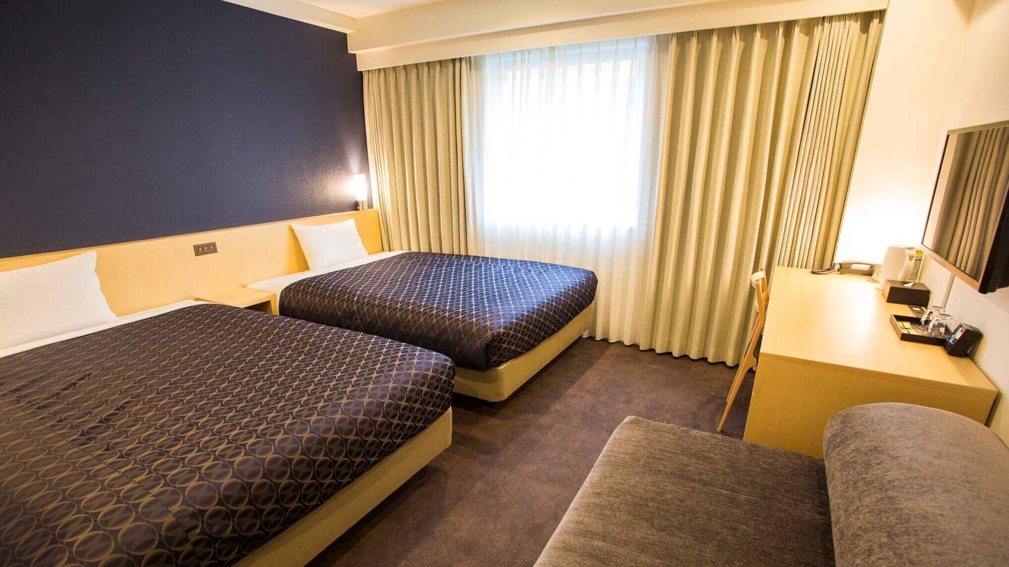 A hotel room with two beds, a desk, a chair, and a large window with curtains.