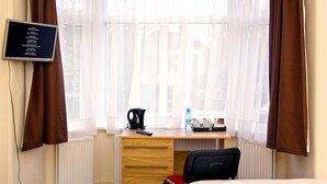 A hotel room with a bay window, a wooden desk, a chair, a black office chair, a blackboard with names, a kettle, a water bottle, and a tray with cups and a tissue box.