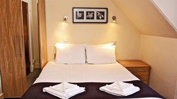 A neatly made bed with white linens and two towels on a dark brown bedspread, a wooden nightstand with a lamp, and a framed picture above the bed.