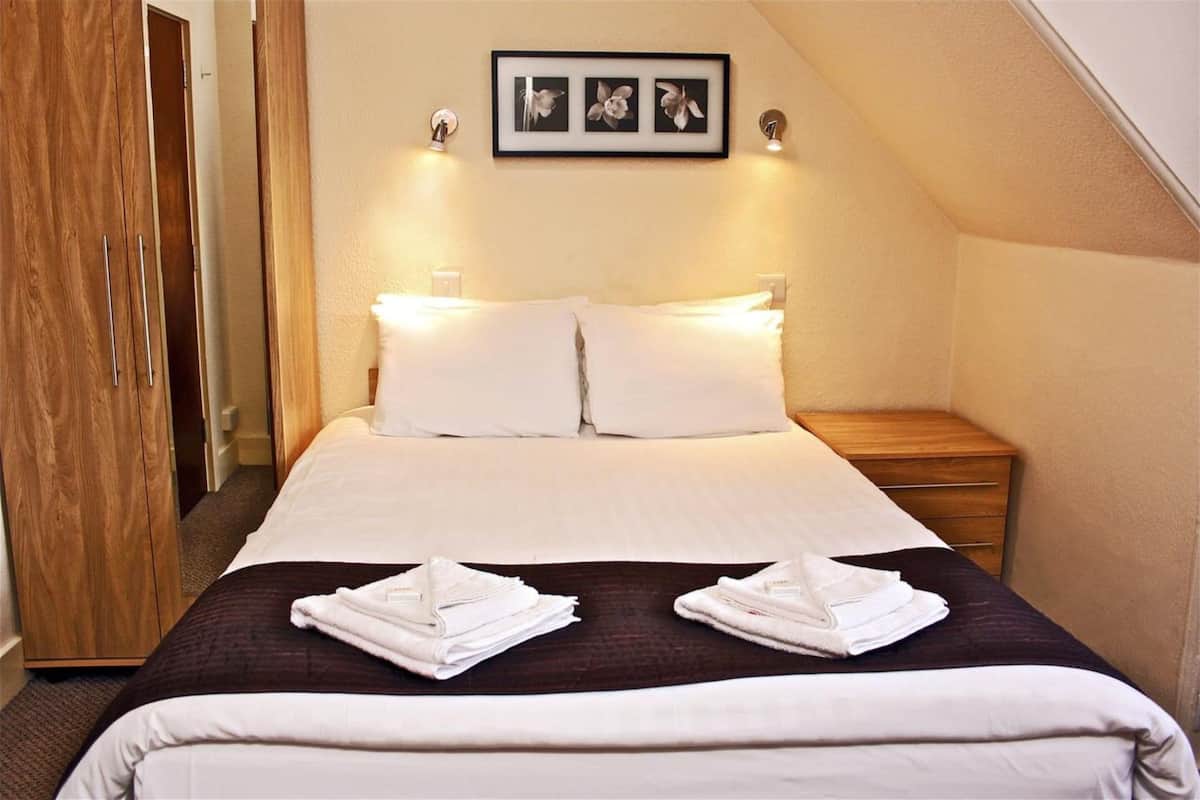 A neatly made bed with white linens and two towels on a dark brown bedspread, a wooden nightstand with a lamp, and a framed picture above the bed.
