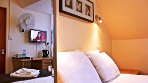 A hotel room with a bed, pillows, a desk with a television, a fan, and a wall-mounted picture.