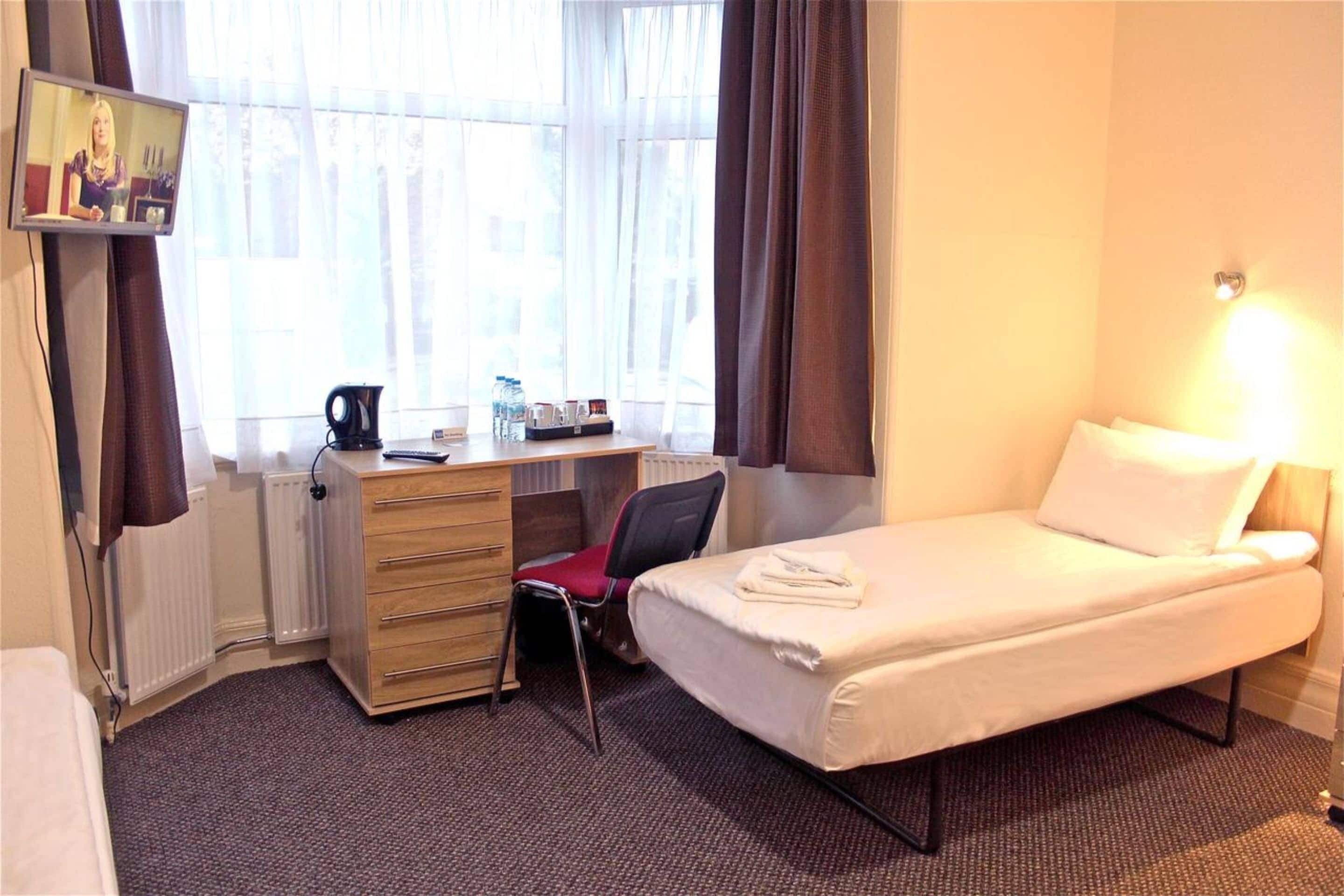 A hotel room with a single bed, a desk with a chair, a television mounted on the wall, and a window with curtains.