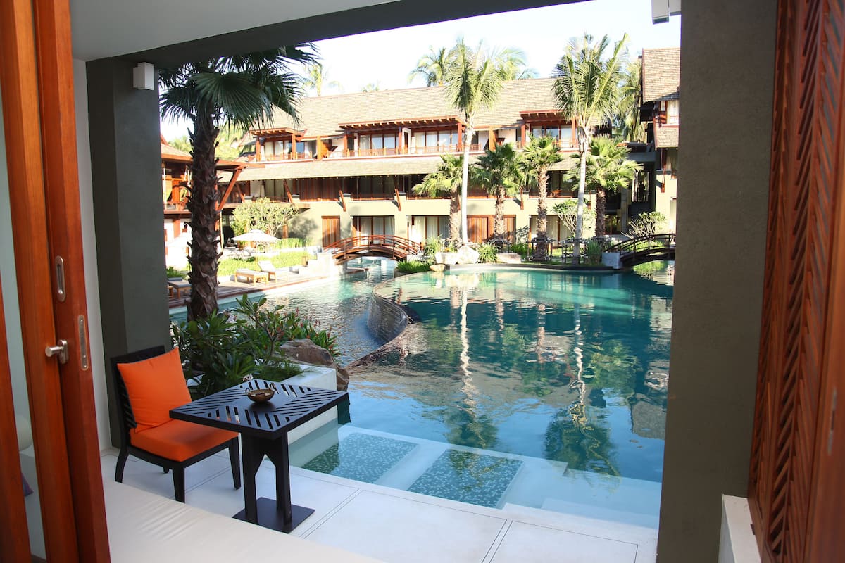 A pool area with a view of a hotel building, a palm tree, and a small bridge.