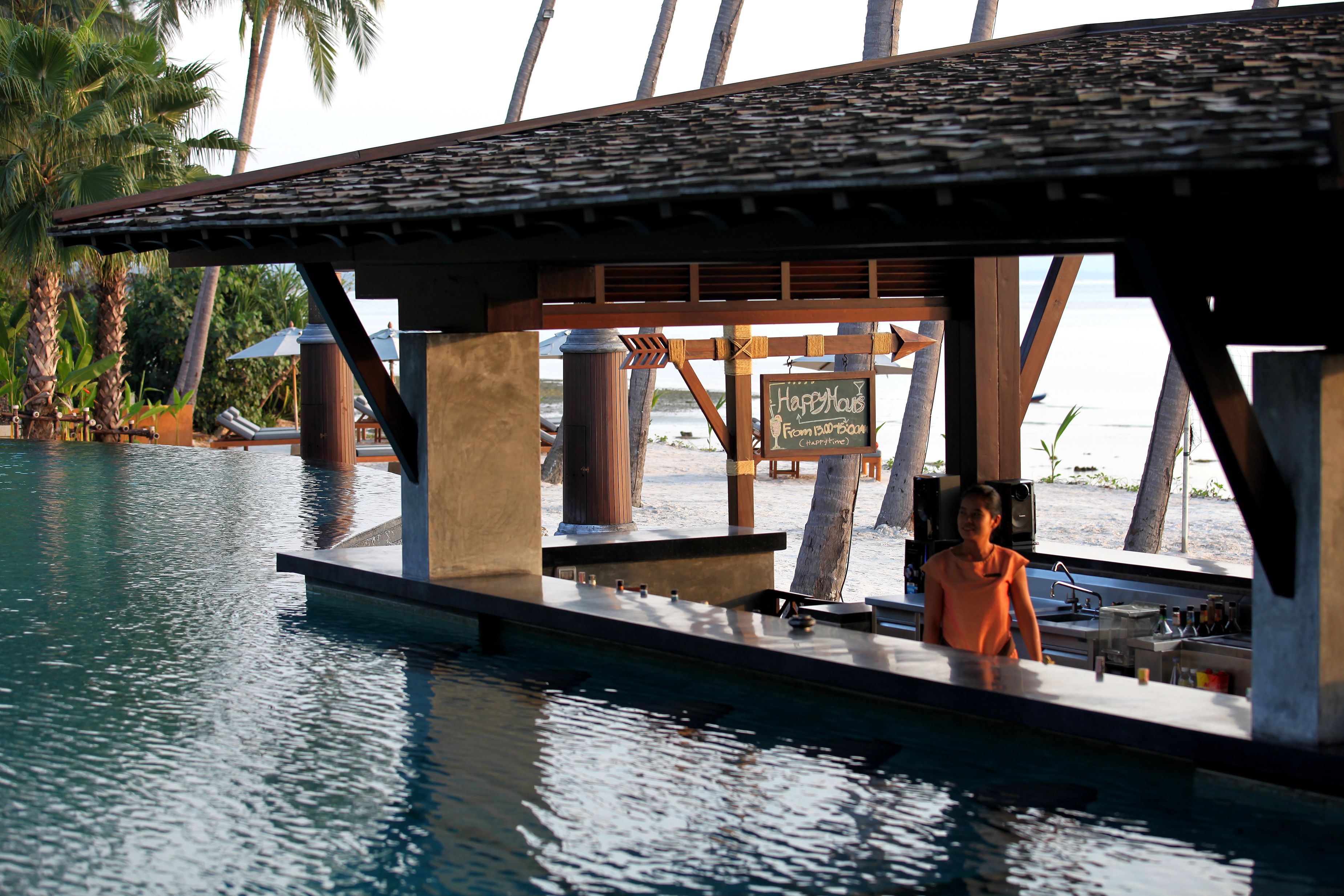 A poolside bar area with a sign reading "Happy Hour," a wooden structure, and a view of the beach.