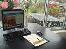 Um monitor de computador exibindo um site de hotel, um teclado, um rato, um bloco de notas com uma caneta e um vaso com flores.