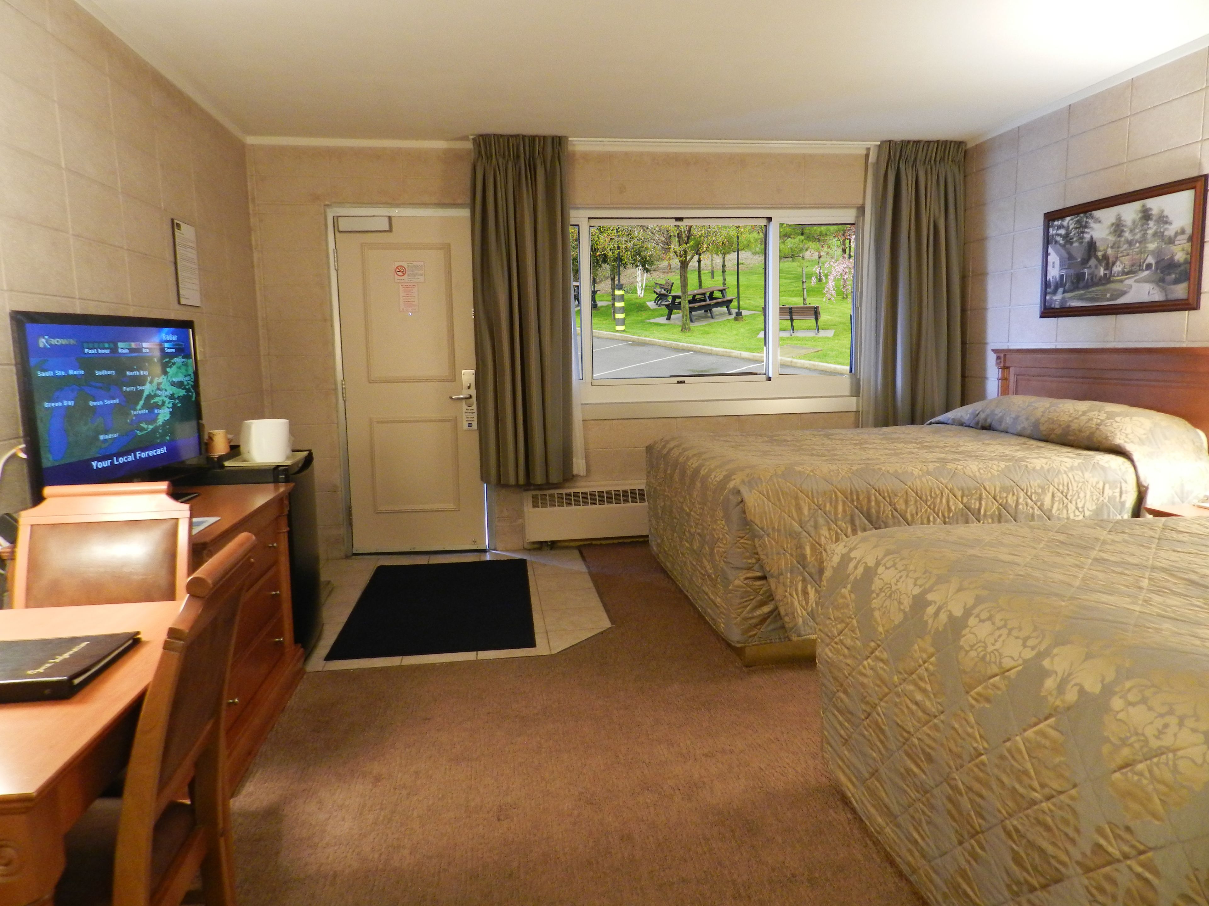 A hotel room with two beds, a desk with a computer, a window with a view of a park, and a painting on the wall.