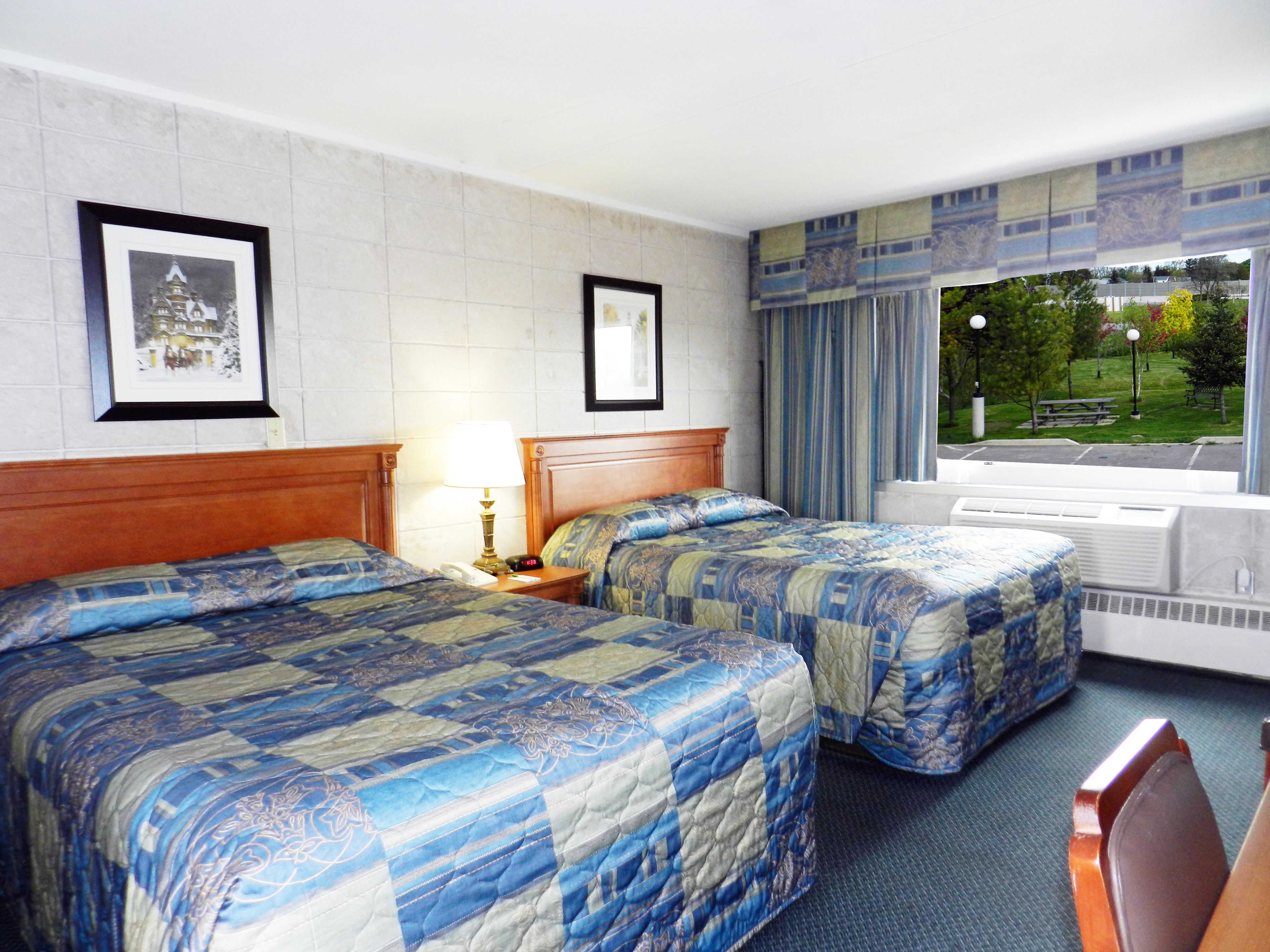 A hotel room with two beds, a nightstand, a lamp, framed pictures on the wall, and a window with a view of a park.