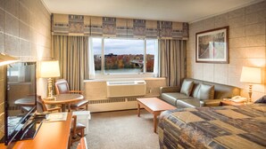 A hotel room with a large bed, a sofa, a desk with a chair, a small table, and a window with a view of trees and a bridge. - Capri Inn (St. Catharines)