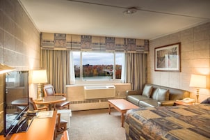 Quarto de hotel com uma cama grande, um sofá, uma escrivaninha com cadeira, uma mesinha e uma janela com vista para árvores e uma ponte.