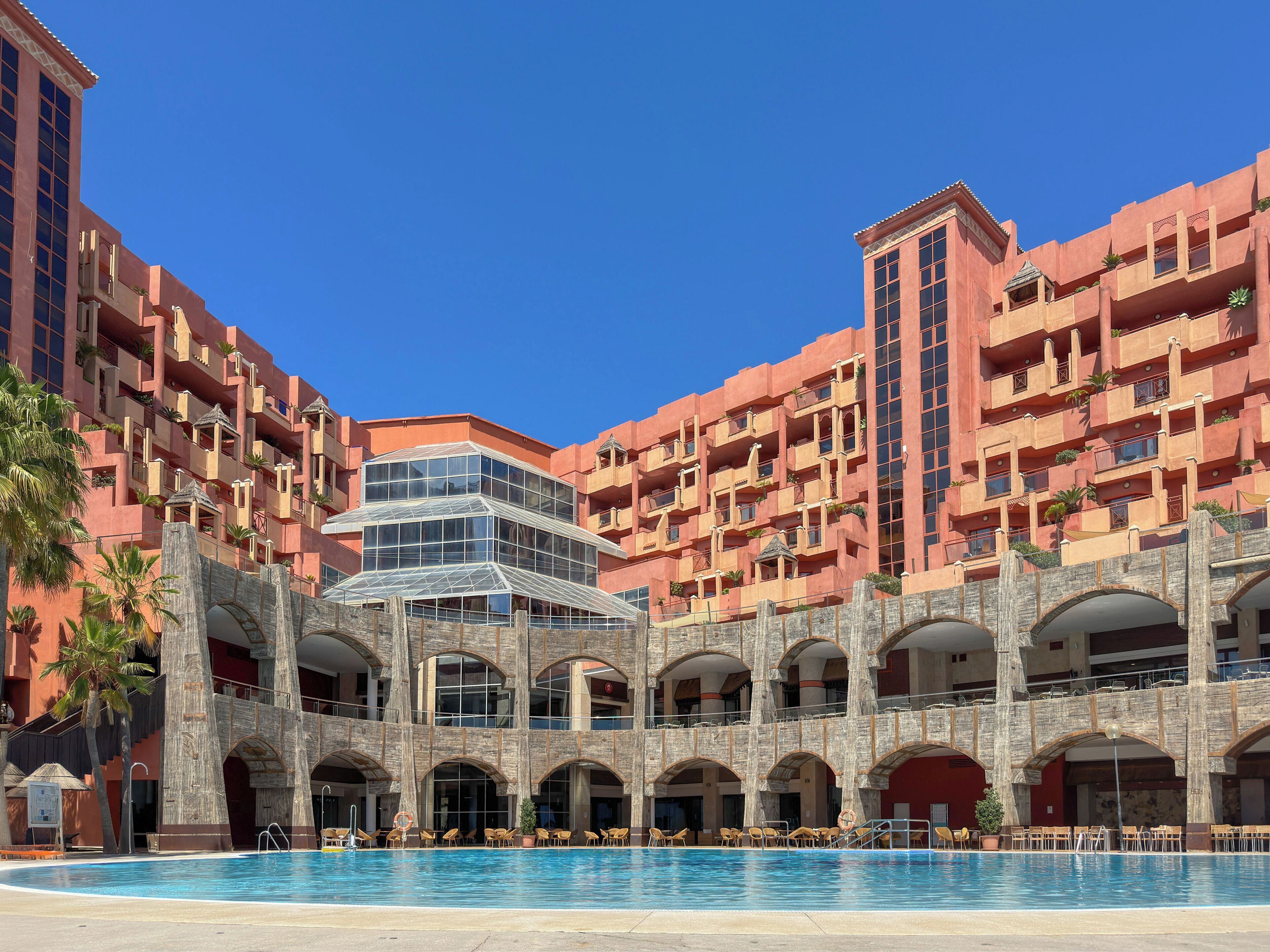 Ett hotell med pool, flera balkonger och en centralt placerad glasstruktur.