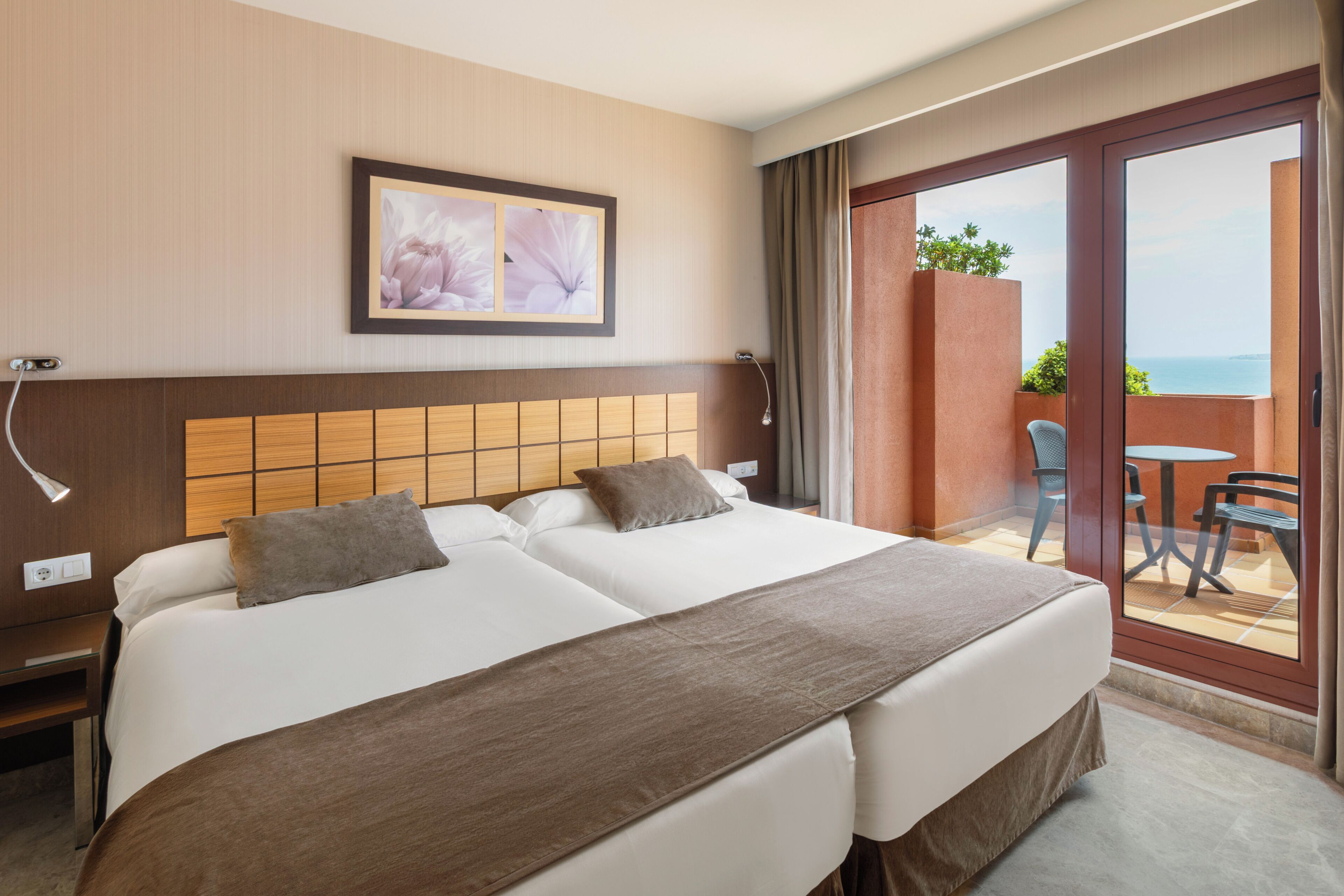 A hotel room with two beds, a wooden headboard, a framed picture on the wall, and a balcony with a table and chairs.