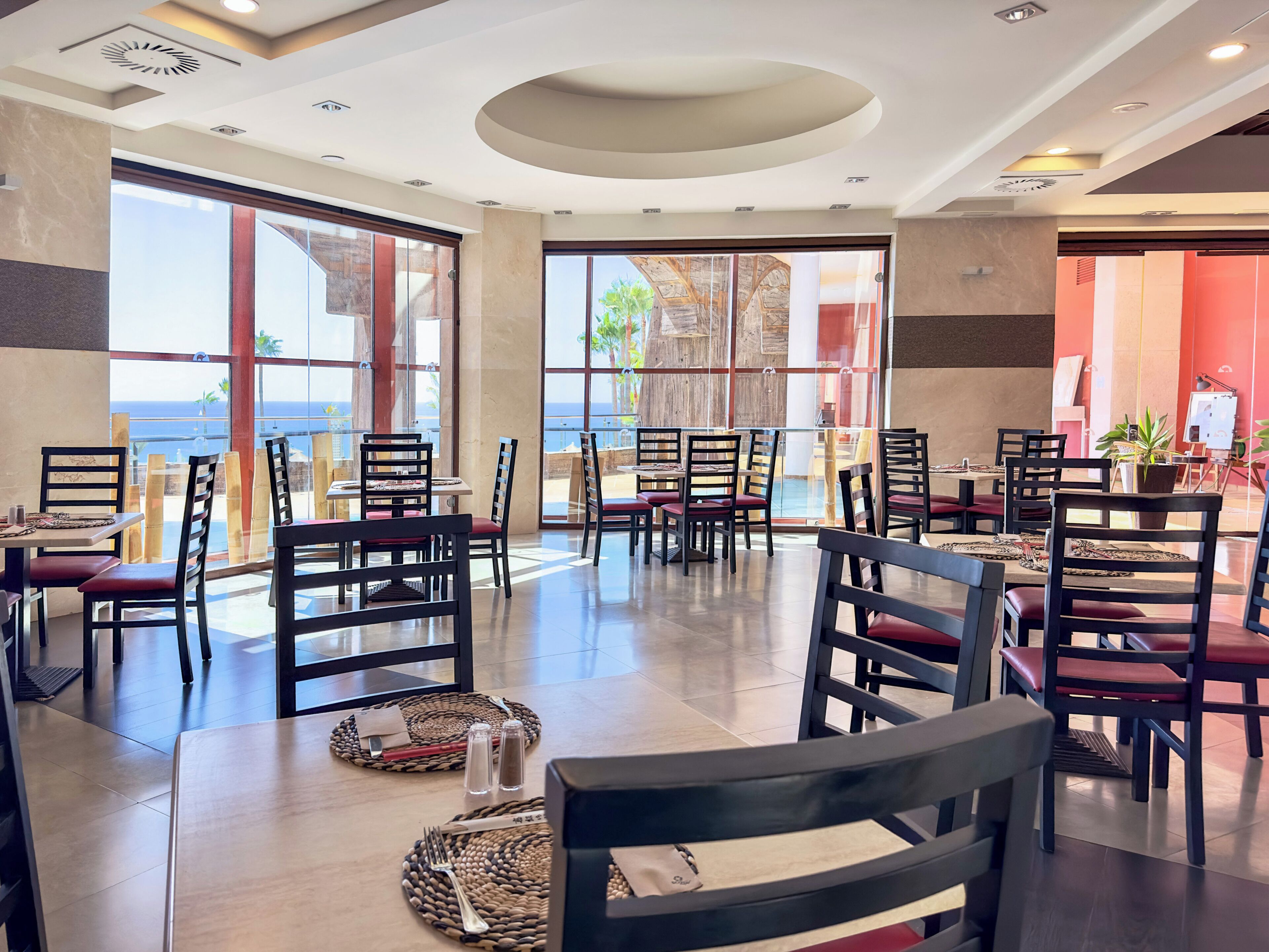 A restaurant with tables and chairs, large windows, and a view of the sea.