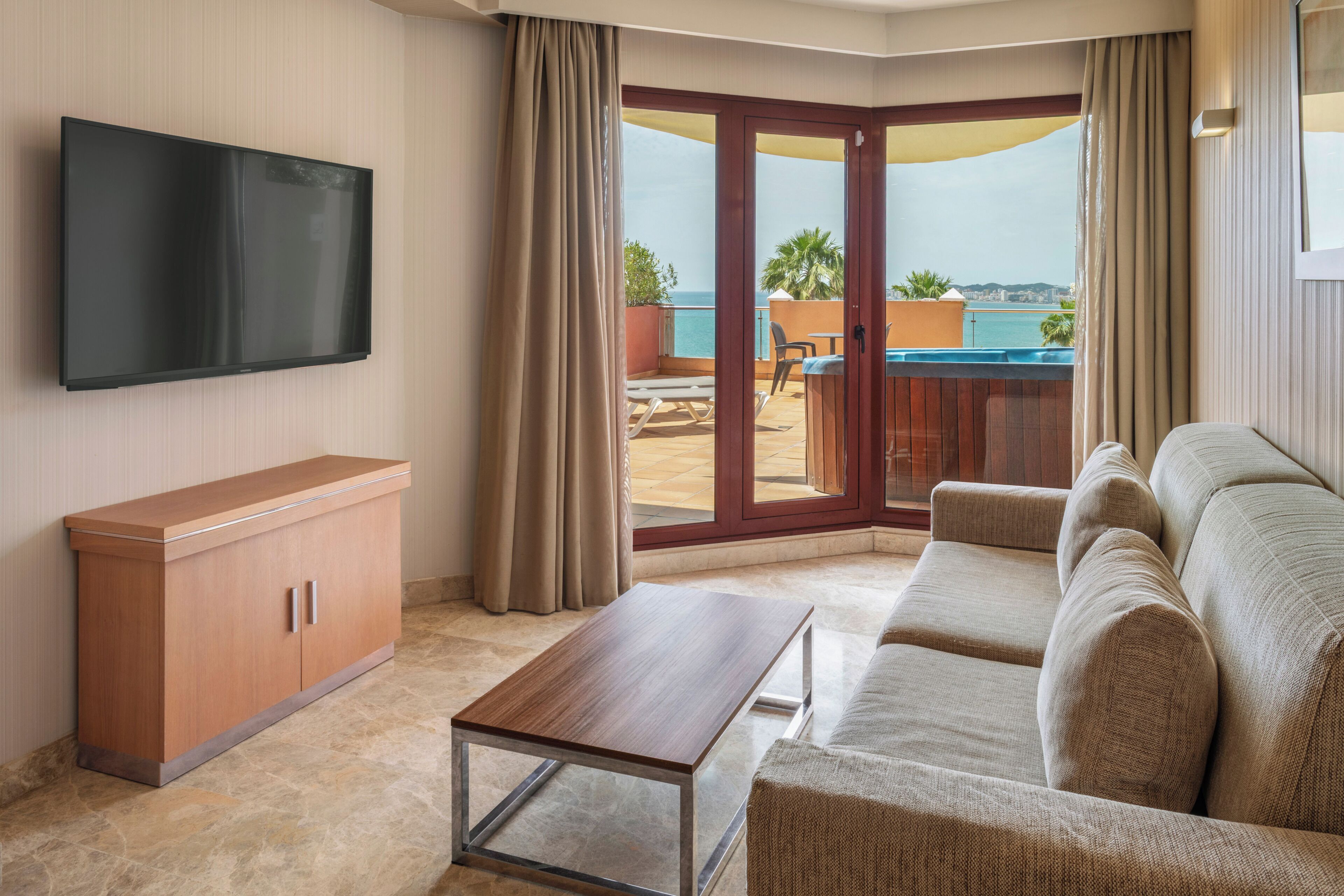 Une chambre d’hôtel équipée d’un téléviseur à écran plat, d’une armoire en bois, d’un canapé, d’une table basse et d’une porte-fenêtre coulissante donnant sur un balcon avec vue sur la mer.